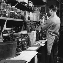 A black and white photo of a man standing at a lab bench, holding up a glass jar