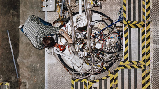 An overhead shot of an engineer working on a metal tank with pipes and cables coming out of it.