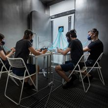 Four study participants in t shirts and shorts sit around a table in a stainless steel chamber. All four are looking at personal electronics and wearing a breathing mask connected to a nearby machine via blue tubing.