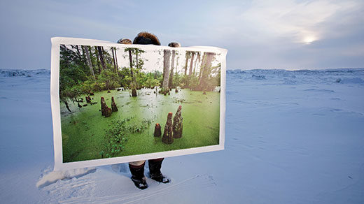 A man standing in the Arctic tundra holds up a large photo of a swamp.
