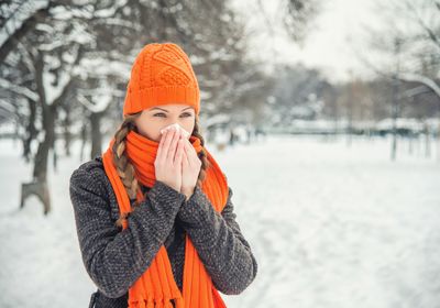 A woman wearing a gray sweater and a bright orange scarf and hat blows her nose vaguely in the direction of the camera. A snowy landscape can be seen behind her.