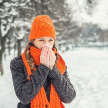 A woman wearing a gray sweater and a bright orange scarf and hat blows her nose vaguely in the direction of the camera. A snowy landscape can be seen behind her.