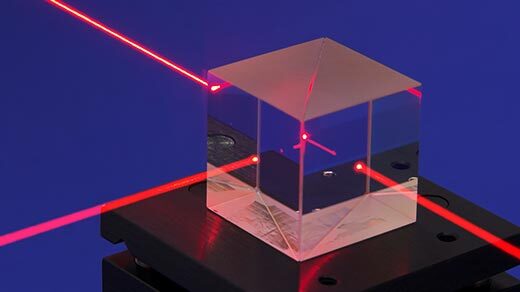 A red laser beam enters a glass cube and splits in two; half of the beam continues straight ahead and the other half shoots out of the glass cube at a right angle.