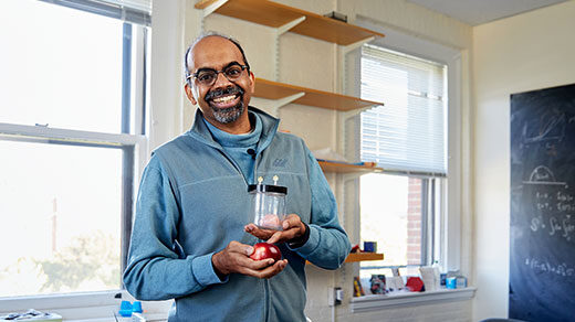 Harvard professor L. Mahadevan stands in a slightly cluttered office, holding an apple and a jar containing a miniature brain