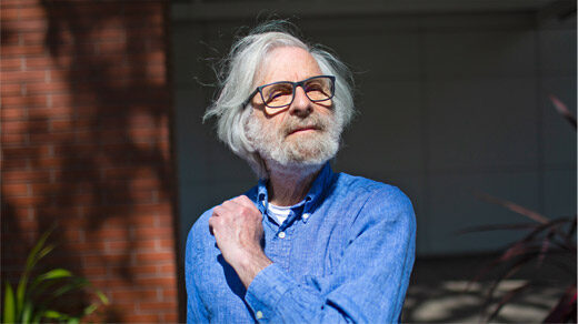 Outdoor photo of Leslie Lamport wearing a blue shirt with his hand up to his shoulder