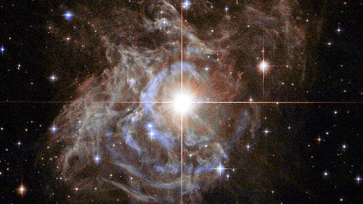 Lede art for "A Radically Conservative Solution for Cosmology’s Biggest Mystery": This Hubble image shows RS Puppis, a type of variable star known as a Cepheid variable. As variable stars go, Cepheids have comparatively long periods— RS Puppis, for example, varies in brightness by almost a factor of five every 40 or so days. RS Puppis is unusual; this variable star is shrouded by thick, dark clouds of dust enabling a phenomenon known as a light echo to be shown with stunning clarity. These Hubble observations show the ethereal object embedded in its dusty environment, set against a dark sky filled with background galaxies.