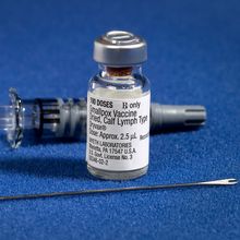 A two-pronged needle, a glass vial of smallpox vaccine, and a syringe sit on a blue surface.