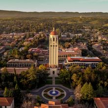 An aerial view of the Stanford University campus. Trees surround several buildings with the bell tower in the center.&nbsp;&nbsp;