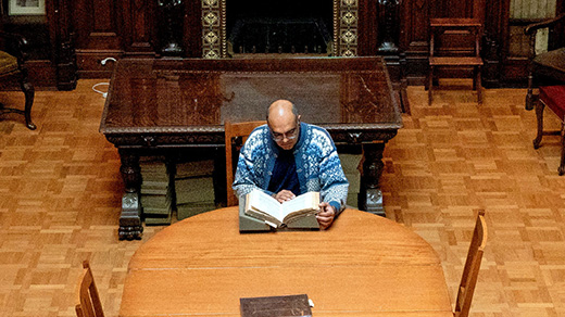 A man sits at a table in the middle of a wood-paneled library, perusing a large book.