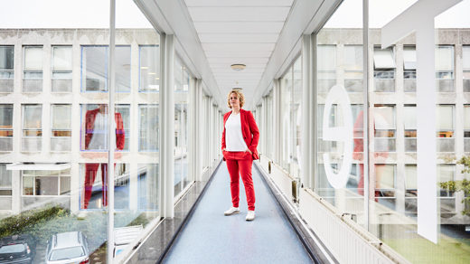 Stephanie Wehner in a red suit standing in a glass-paneled corridor at Delft University of Technology in the Netherlands. Her reflection appears in the glass to the right and left.